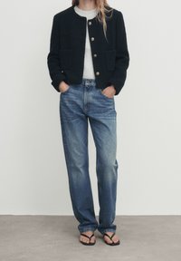 Svart, texturerad kort jacka med guldknappar, grå topp under, tillsammans med blå högmidjade denimjeans och svarta flip-flops.