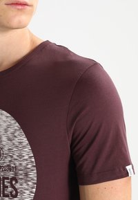 Närbild av en person som bär en vinröd t-skjorta med korta ärmar, med en delvis tryckt design och en liten "CORE"-etikett på ärmen.