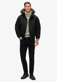 Czarna puchowa kurtka z kapturem wyłożonym futrem, z zamkiem z przodu, dwoma bocznymi kieszeniami, noszona na jasnoszarym swetrze i czarnych spodniach.