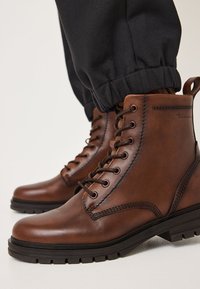 Stivali ankle in pelle marrone con punta rotonda, design con lacci, cuciture laterali e suola in gomma testurizzata. I pantaloni presentano polsini elasticizzati.