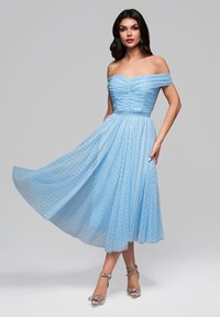 Ljusblå off-shoulder klänning med rynkad bodice, flödande veckad kjol och prickig textur. Matchad med silverfärgade sandaler med remmar.