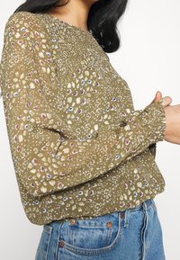 Langarmbluse aus olivgrünem Stoff mit einem verstreuten Blumenmuster in Creme, Rot und Blau. Elastische Bündchen mit Raffung.