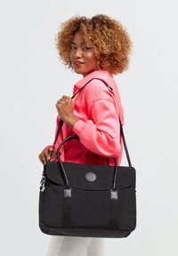 Schwarze Laptoptasche mit zwei Griffen und einem abnehmbaren Trageriemen. Verfügt über einen Lederakzent, ein Logo und eine glatte Textur. Wird von einer Person mit einer leuchtend pinken Jacke getragen.