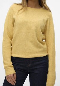 Kvinna iklädd en mjuk, gul tröja med långa ärmar och mörkblå jeans, stående mot en enkel ljus bakgrund.