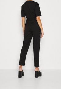 Mujer vestida con un mono negro con cinturón y sandalias de plataforma negras, de pie contra un fondo blanco liso, de espaldas.
