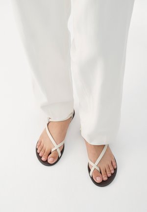 Világosszürke, csatolt szandál lapos fekete talppal; keresztpántos kialakítás és fém csat található az oldalon; fehér nadrággal viselve.