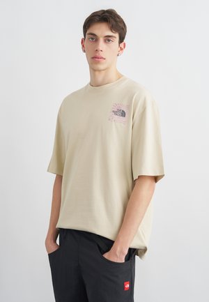 Jeune homme portant un t-shirt beige oversize de The North Face et un pantalon noir, les mains dans les poches, debout devant un fond clair uni.