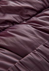 Burgunderfarvet quiltet dyne lavet af glat, shiny stof med synlige sømme og bløde, fyldige sektioner, der skaber et struktureret udseende.