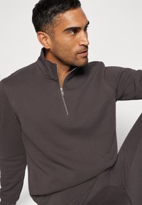Quarter-zip genser i mørkegrått stoff, med ribbestruktur, stående krage og detaljer i sølvglidelås, kombinert med matchende bukser.