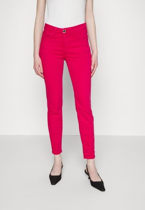 Femme portant un pantalon slim rose vif, un haut blanc sans manches et des chaussures slingback noires à bouts pointus, debout devant un fond neutre.