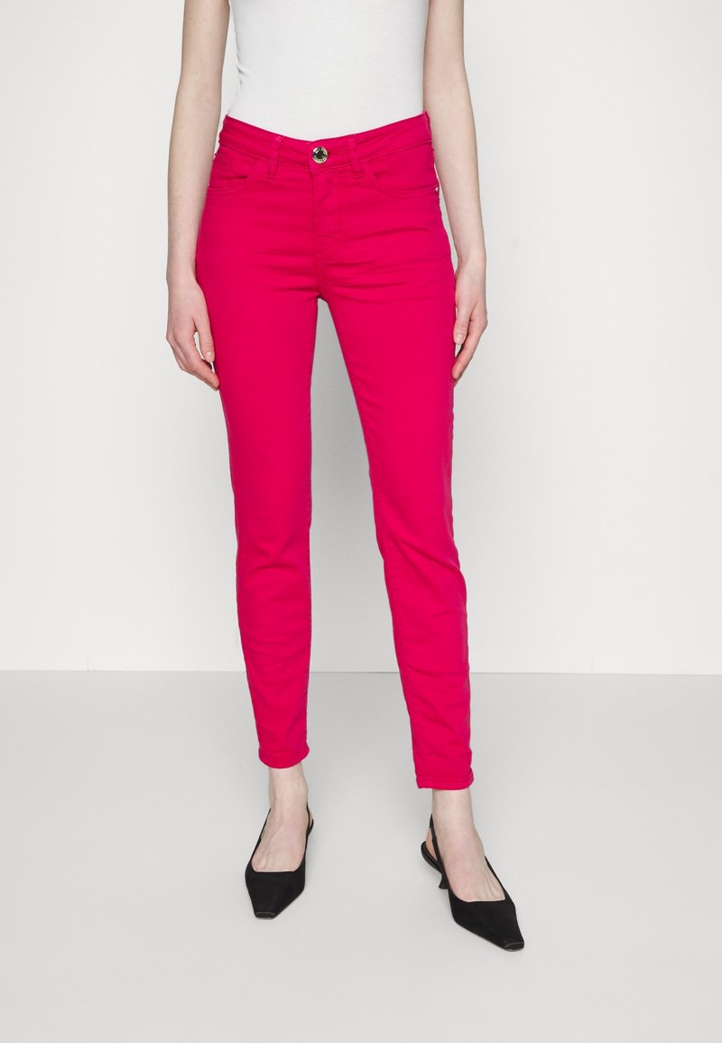Femme portant un pantalon slim rose vif, un haut blanc sans manches et des chaussures slingback noires à bouts pointus, debout devant un fond neutre.