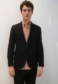 Sacou negru croit, purtat peste o cămașă cu un model geometric în nuanțe de portocaliu și crem. Material neted, cu o croială modernă.
