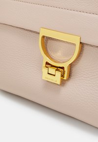 Sac en cuir rose clair avec un fermoir en métal doré bien en vue, comportant une découpe circulaire et une finition texturée. Nom de la marque embossé sous le fermoir.