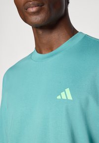 Camiseta turquoise hecha de algodón suave, con cuello redondo y un logo verde claro con tres rayas diagonales en el lado izquierdo del pecho.