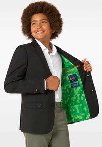 Zēns ar afro frizūru, tērpies melnā žaketē ar dzīvespriecīgu zaļu rakstu oderi, baltā kreklā un olīvzaļās biksēs, smaida, turot žaketi vaļā.