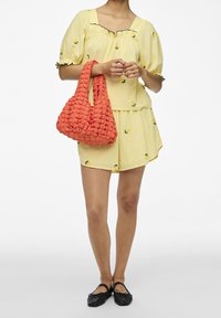 Žlutá bavlněná blůza s nadýchanými rukávy a výšivkou citronu, v kombinaci s odpovídajícími kraťasy. Drží texturovanou oranžovou tašku s tkaným designem.
