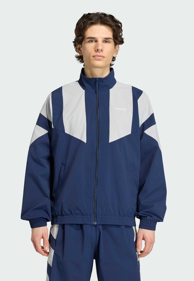 Ung mand iført en mørkeblå og lysegrå Adidas træningsjakke og matchende bukser, stående med hænder afslappet langs siden.