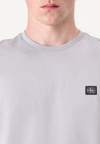 Pelēks īsām piedurknēm T-krekls, kas izgatavots no kokvilnas, ar apaļu kakla izgriezumu un mazu melnu Calvin Klein logotipa uzšuvumu uz kreisās krūts.