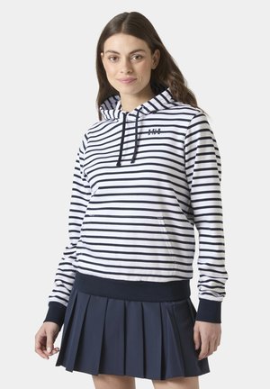 Mujer joven que lleva una sudadera con capucha blanca y azul marino a rayas con un bolsillo delantero y una falda plisada azul marino, de pie frente a un fondo liso.