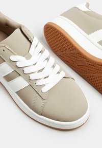 Beżowe sneakersy z białymi akcentami, gładka syntetyczna cholewka, gumowa podeszwa i białe sznurówki. Ma trzy białe paski po boku.