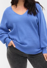 Blauer Strickpullover mit V-Ausschnitt und lässiger Passform. Mit langen Ärmeln und geripptem Material. Kombiniert mit dunkelgrauen Jeans.