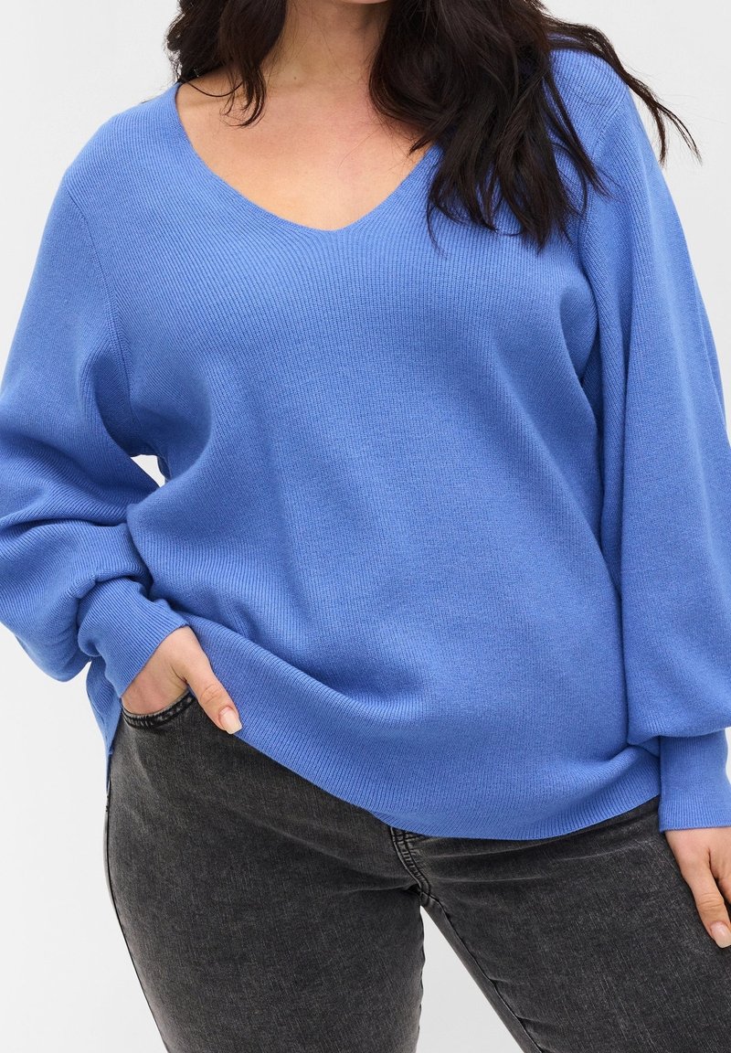 Blauer Strickpullover mit V-Ausschnitt und lässiger Passform. Mit langen Ärmeln und geripptem Material. Kombiniert mit dunkelgrauen Jeans.