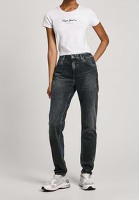 Vit t-shirt med svart logo, parat med högmidjade mörkgrå jeans. Modellen bär vita och grå atletiska sneakers.