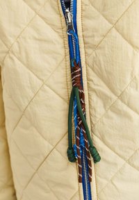 Prošívaná béžová bunda s modrým zipem. Klika zipu je vyrobena z propletených hnědých a zelených šňůrek.