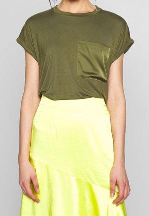 Olivgrünes T-Shirt mit hochgekrempeltem Ärmel-Design und großer Satin-Tasche. Kombiniert mit einem glänzenden, hellgelben asymmetrischen Rock.