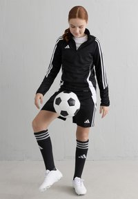 Melnas sporta treniņtērps ar baltajiem akcentiem, melni šorti, svītrainas augstas zeķes un baltas futbola kurpes. Spēlētājs līdzsvaro melnbaltu futbola bumbu.