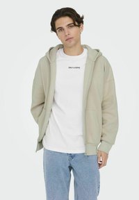 Lichtgroene zip-up hoodie met een zachte textuur, gecombineerd met een wit t-shirt met zwarte letters. Casual blauwe denim jeans maken de look compleet.
