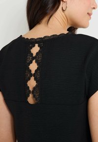 Blusa negra con detalle de encaje en la espalda y bordes en forma de ondas. Textura suave, mangas cortas y escote redondeado.