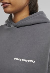 Grijze capuchontrui met een zachte textuur en een witte "PROHIBITED" logo aan de voorkant. Close-upweergave toont de details van de stof en het ontwerp.