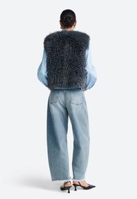 Grijze nepbont-vest met een ronde halslijn, gedragen over een lichtblauw overhemd, gecombineerd met lichtgekleurde high-waisted spijkerbroek en zwarte schoenen met hak.