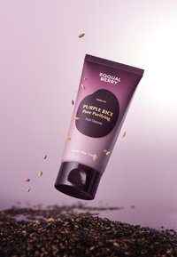 Fioletowy żel oczyszczający w miękkiej fioletowej tubie, z czarną etykietą zawierającą nazwę produktu i informacje, spoczywający na poduszce ciemnych ziaren.