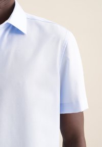 Camicetta azzurra a maniche corte con una texture liscia e compatta. Presenta un colletto classico e cuciture pulite lungo le cuciture delle spalle.