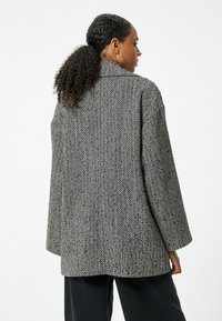 Płaszcz w wzór houndstooth w odcieniach czerni i bieli, w oversize'owym kroju, z wyraźnym kołnierzem i długimi rękawami, o luźnym fasonie i teksturowanej tkaninie.