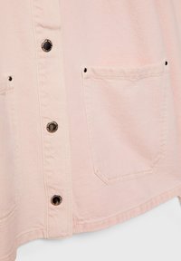 Camisa de mezclilla rosa claro con botones de presión negros, un bolsillo en el pecho y un dobladillo ligeramente curvado con tejido texturizado.