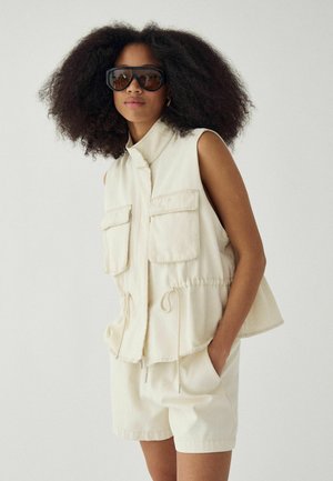 Žena s objemnými kučeravými vlasmi, ktorá má na sebe veľké čierne slnečné okuliare, bezrukávovú béžovú bundu so sťahovacou šnúrkou a zodpovedajúce šortky, stojaca s rukami v vreckách.