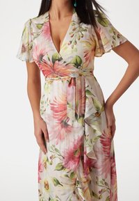 Mujer vestida con un vestido envolvente de flores con escote en V, mangas abullonadas y cinta en la cintura, con patrones de flores en rosa, amarillo y verde.