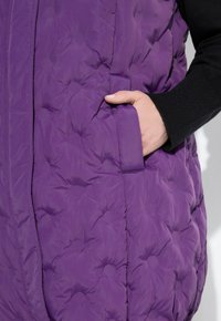 Hand in der Tasche eines lila gepolsterten Mantels, der über einem schwarzen gerippten Ärmelkleidungsstück vor weißem Hintergrund getragen wird.