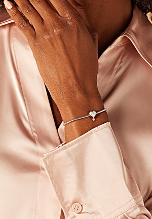 Zilveren armband met een hartbedel en een druppeldetail, gedragen om een hand tegen een zacht roze satijnen blouse. Heeft een slank, flexibel ontwerp.