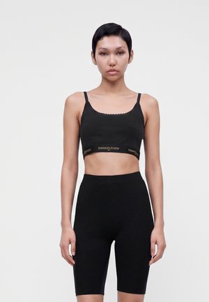 Jeune femme aux cheveux courts portant un crop top noir à fines bretelles et un short de cycliste taille haute assorti sur un fond blanc uni.