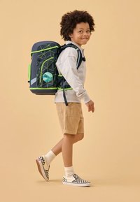 Lo zaino presenta un corpo grigio scuro con accenti verdi, motivi geometrici e un logo rotondo. Il bambino indossa una felpa grigio chiaro e pantaloncini color sabbia.