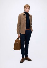 Jeune homme portant une veste camel, un col roulé noir, un pantalon bleu marine et des bottes marron, tenant un sac en cuir marron dans sa main droite.