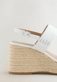 Witte wedge sandaal met een gevlochten jute zool, voorzien van een gladde bovenste band en een verstelbare zilveren gesp. Ronde te ontwerpen.