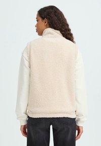 Gilet di pile beige con colletto alto, tessuto testurizzato e tasche laterali, indossato sopra una camicia bianca a maniche lunghe e jeans scuri.