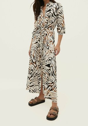 Vrouw die een lange, zebraprint, jurk met knopen draagt, met opgerolde mouwen en bruine sandalen met gesp, staand tegen een effen achtergrond.