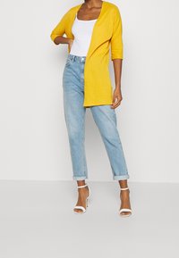 Gul ribbad kavaj med trekvartslånga ärmar, tillsammans med ljusblå uppvikta jeans och vita klacksandaler.