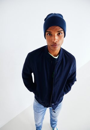 Chaqueta bomber azul marina con cremallera y puños acanalados, combinada con un gorro de punto azul oscuro y jeans azul claro. Fondo sencillo.